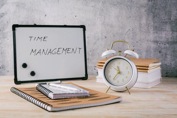 Ein weißer Wecker, Notizhefte und ein Whiteboard mit der Aufschrift „TIME MANAGEMENT“ - Zeitmanagement - stehen auf einem hellen Holztisch vor einer grauen Betonwand.