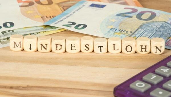 Die Würfelreihe Mindestlohn liegt auf einem Holztisch, im Hintergrund sind Euro-Geldscheine und rechts eine violette Rechenmaschine angeschnitten.