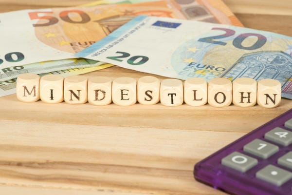 Die Würfelreihe Mindestlohn in Deutschland liegt auf einem Holztisch, im Hintergrund sind Euro-Geldscheine und rechts eine violette Rechenmaschine angeschnitten.