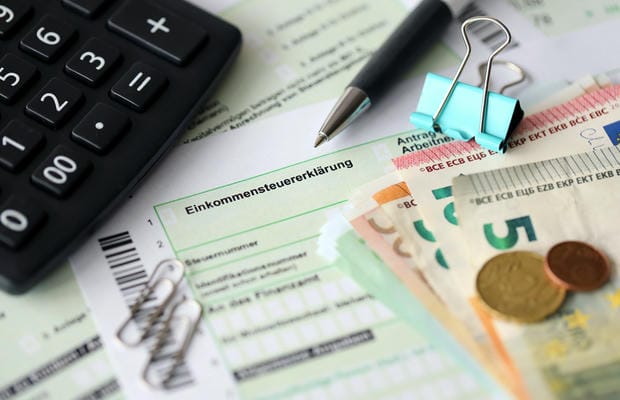 Ein deutsches Formular zur Einkommensteuererklärung liegt bereit, umgeben von einem Taschenrechner, Euro-Geldscheinen und Münzen sowie einem Stift für die Berechnung des Netto-Verdienstes.
