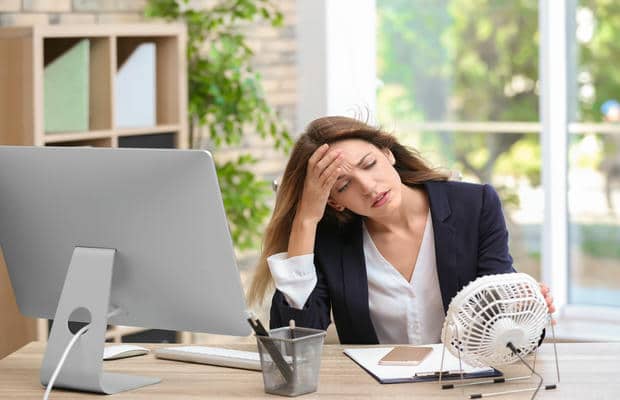Eine erschöpfte Frau sitzt wegen der Hitze am Büroschreibtisch und richtet einen kleinen Tischventilator auf ihr Gesicht.
